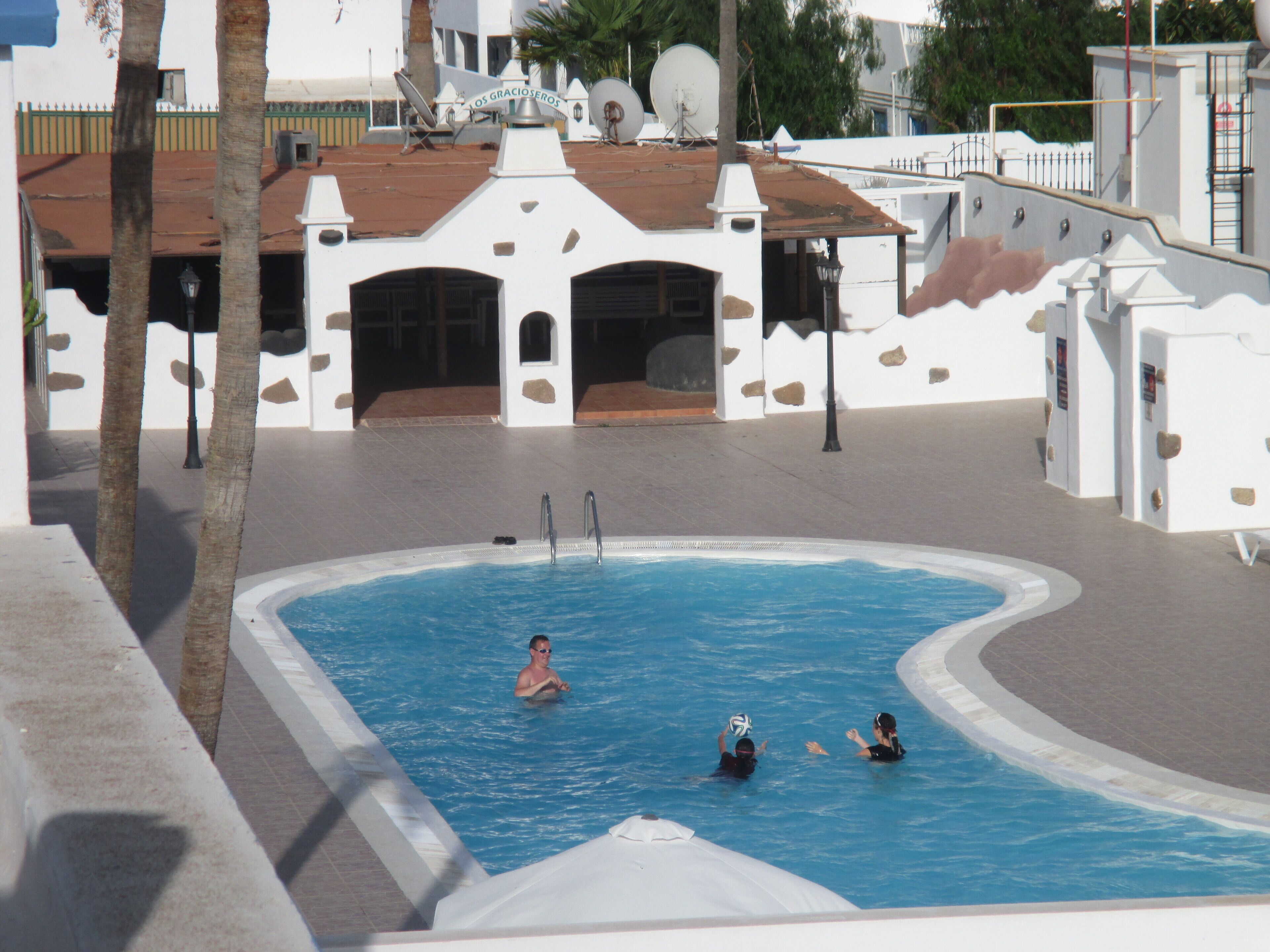 A swimming pool with a tiled deck, a building with a white facade and stone accents, and palm trees in the background.