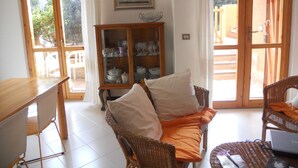 Ein Wohnzimmer mit Holztisch, Korbstühlen, einer Couch mit Kissen, einer Vitrine mit Glastüren und einem Gemälde an der Wand.
