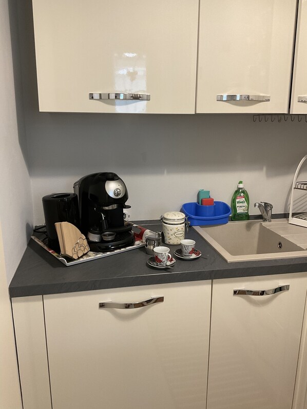 A kitchen with a coffee maker, cups, a kettle, and a sink.