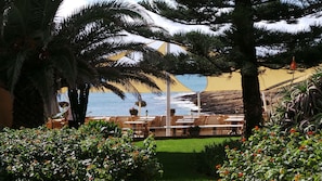 Un espace extérieur aménagé avec des tables et des chaises, entouré d’une végétation luxuriante et de palmiers, offrant une vue imprenable sur la côte.