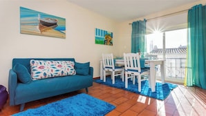 A living room with a blue sofa, a dining table with white chairs, and a balcony with a view. - WAKE UP TO THE SOUND OF THE SEA T1 CENTRAL-AIR CONDITIONING(FREE WIFI) 3' BEACH (Luz)