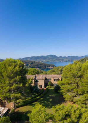 Una villa con tejado de teja anaranjada, rodeada de abundante vegetación y vistas a un lago y colinas distantes.