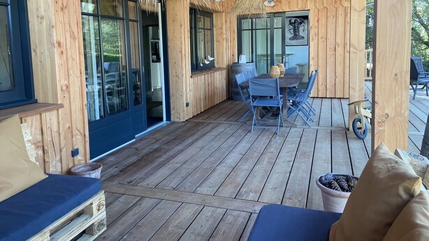 Eine Holzterrasse mit Tisch und Stühlen, einer Hängelampe und einem großen Fenster.