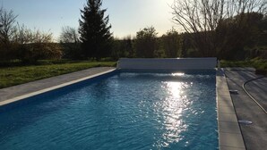 Una piscina rectangular con agua azul cristalina, rodeada por una terraza de concreto y un área verde con árboles.