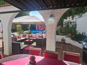 Une terrasse extérieure dotée d’un espace salon couvert, d’une piscine et de plantes en pot.