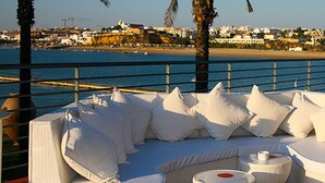 A seaside terrace with white outdoor furniture, including a sofa and chairs, overlooking a beach and a historic fort.