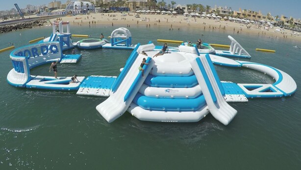 An inflatable water park with slides and play structures in the water, surrounded by a beach and buildings in the background.