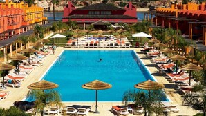 A large swimming pool with sun loungers and umbrellas, surrounded by palm trees and buildings with red and orange facades.