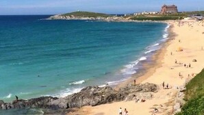 Una spiaggia costiera con un ampio edificio sullo sfondo, cielo sereno e riva rocciosa.