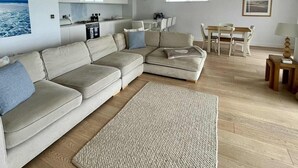Un soggiorno moderno con un ampio divano ad angolo, un'area pranzo con un tavolo e sedie in legno, e una cucina con mobili bianchi e un piano cottura.