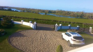 A white car parked in a gravel driveway with a view of a lake and fields. - Architect Designed Stunning House, In Quiet And Peaceful Stunning Location  (Tenby. Pembrokeshire)