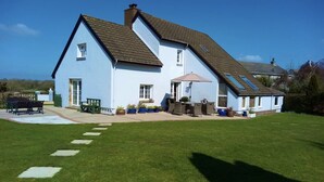 A two-story house with a sloped roof, a patio area with outdoor furniture, and a well-maintained lawn. - Architect Designed Stunning House, In Quiet And Peaceful Stunning Location  (Tenby. Pembrokeshire)