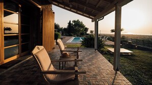 A covered patio with two lounge chairs, a small table, and a view of a swimming pool and landscape. - Luxury Prinate Villa in Aegina (Aegina)