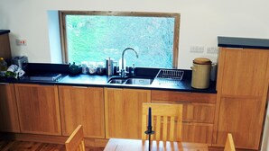 Una mesa de comedor de madera con cuatro sillas, una ventana que da a zonas verdes y una cocina con armarios de madera y un lavamanos.