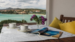 Une table donnant sur un paysage côtier, un vase rempli de fleurs violettes et un magazine posé sur une nappe.