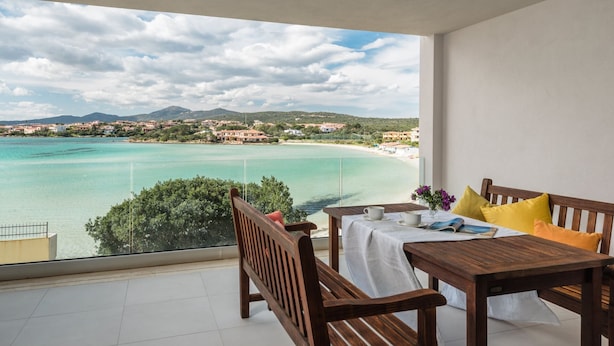 Un balcon avec une table et des chaises en bois, une nappe blanche et une vue sur une ville côtière et une mer turquoise.