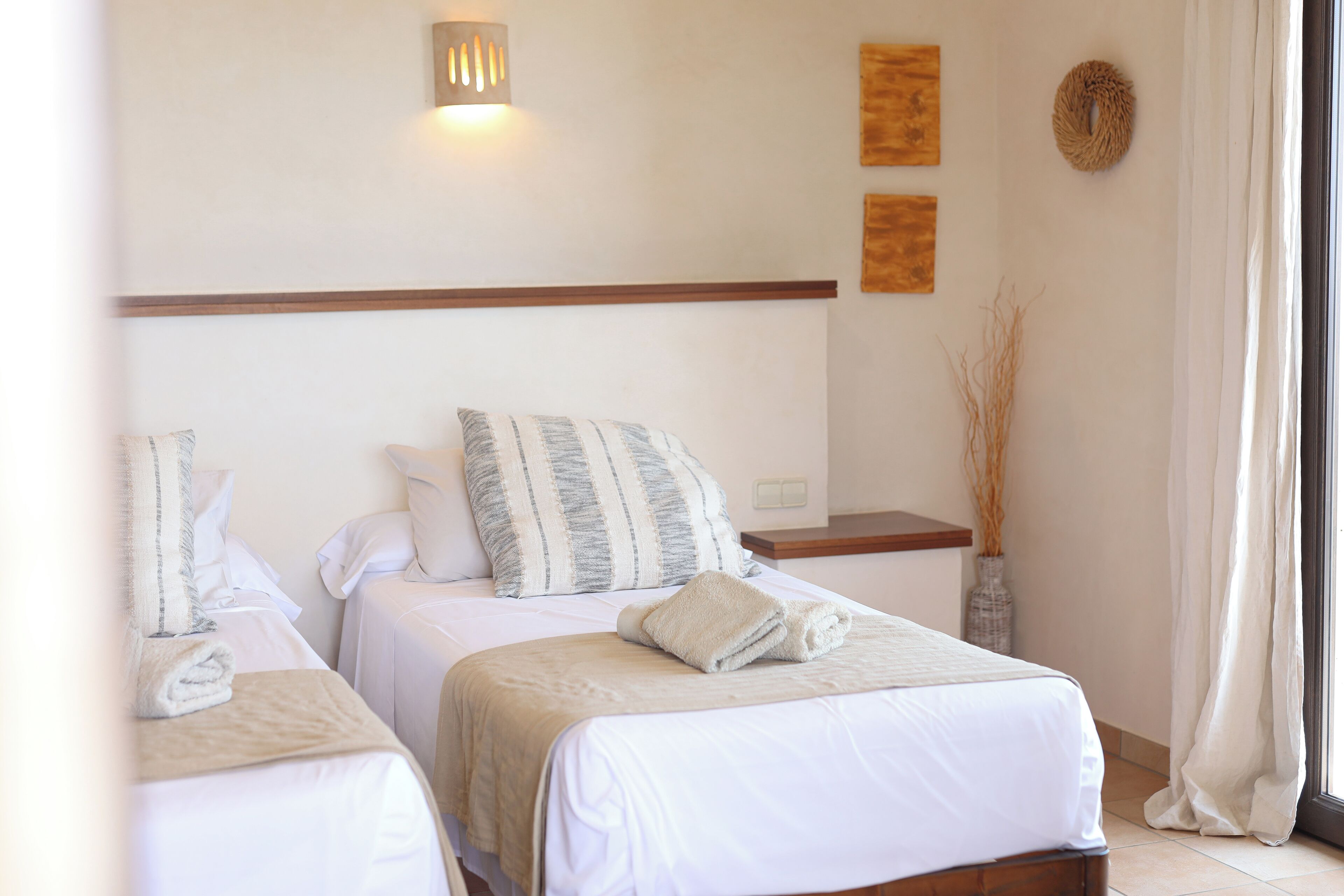 Habitación de hotel con dos camas, una mesita de noche, un jarrón con plantas secas y una ventana con cortinas.