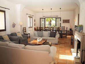 Amplia sala de estar con chimenea, comedor con mobiliario de madera y vista a palmeras a través de grandes ventanales.