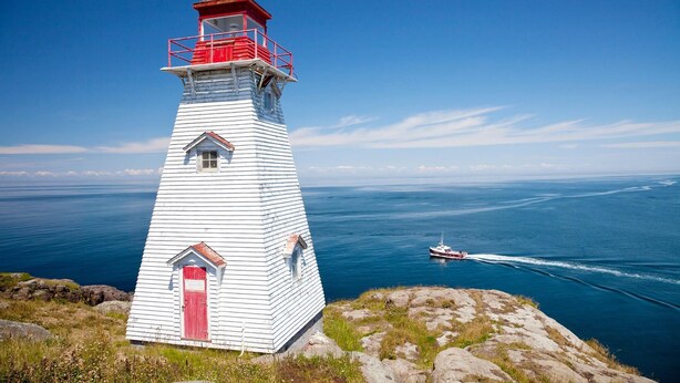 A lighthouse with a red top and white body, situated on a rocky cliff overlooking a calm sea with a boat in the distance. - Home from Home -  Cosy Cottage in Whale Watching Country (Little River)