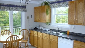 A kitchen with wooden cabinets, a white dishwasher, a granite countertop, and a dining area with wooden chairs. - Home from Home -  Cosy Cottage in Whale Watching Country (Little River)