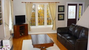 A living room with a leather sofa, a wooden coffee table, a TV on a wooden stand, and a window with curtains. - Home from Home -  Cosy Cottage in Whale Watching Country (Little River)