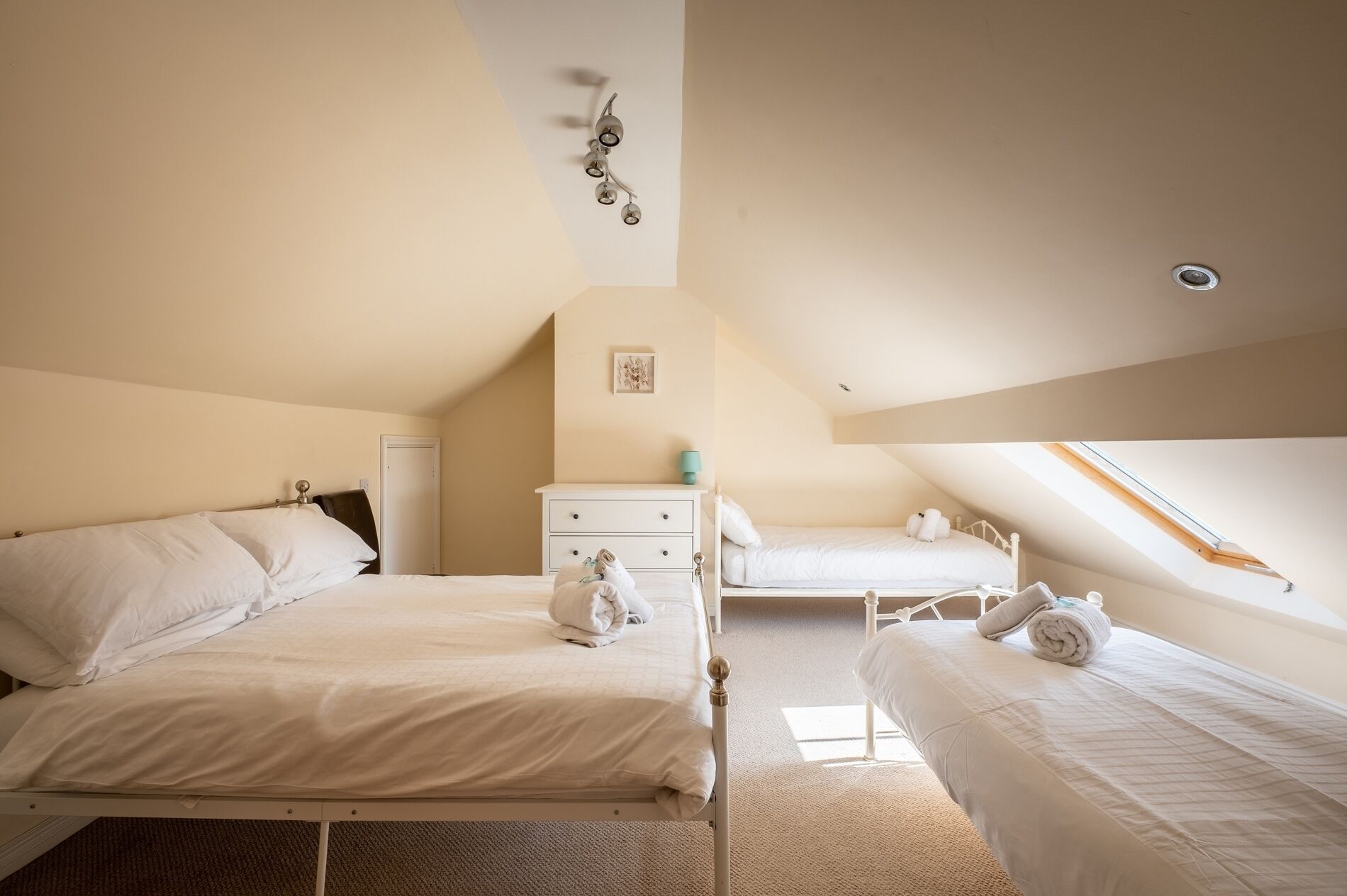 A bedroom with two beds, a white dresser, a small table, and a wall-mounted light fixture.