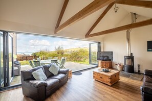 En rummelig stue med en lædersofa, et træbord og et fjernsyn. En stor skydedør fører ud til en terrasse med havemøbler og en smuk udsigt.