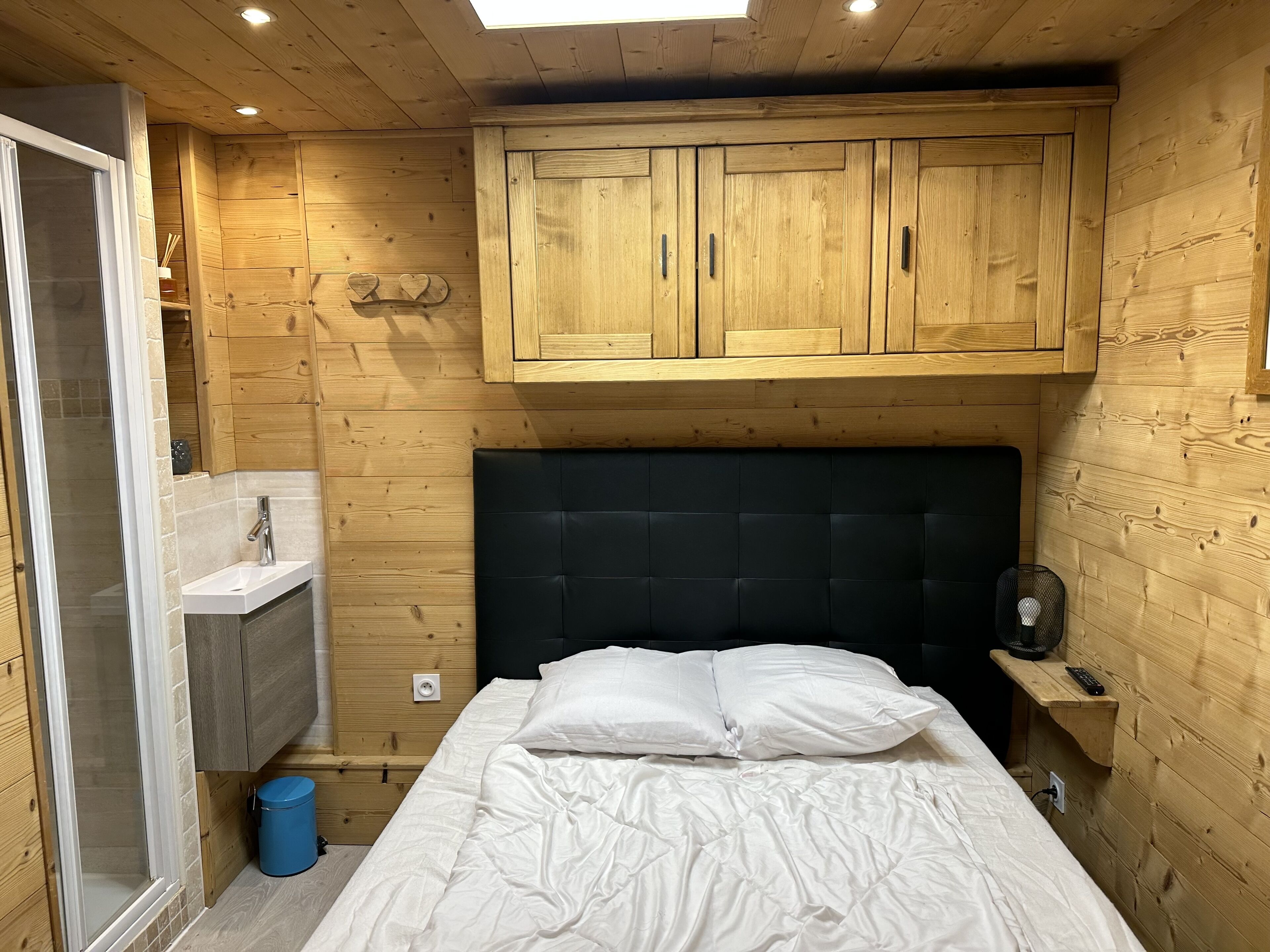 Ein gemütliches Schlafzimmer mit Holzbalkendecke, einem schwarzen Kopfende, einem weißen Bett, einem kleinen Nachttisch mit Lampe und einem Holzschrank.