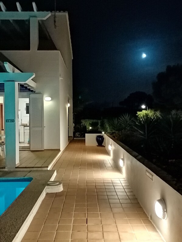 A nighttime view of a modern building with a tiled patio, a pool, and a well-lit pathway leading to the entrance.