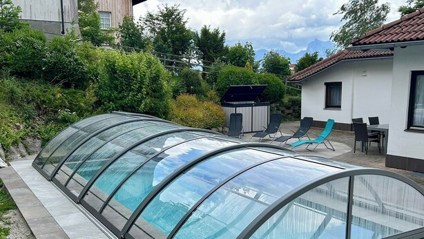 A covered swimming pool with a glass roof, surrounded by a patio area with chairs and a small outdoor kitchen. - Hillside villa with private pool (Altmünster)