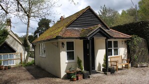 A small, single-story house with a sloped roof, a front porch with a bench, and several potted plants. - Amberfold Lodge, Heyshott near Midhurst (Midhurst)