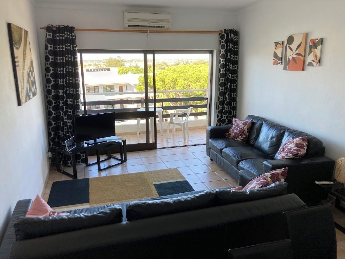 A living room with a black leather sofa, a TV, and a balcony with a view of a building named LEZIRIA.