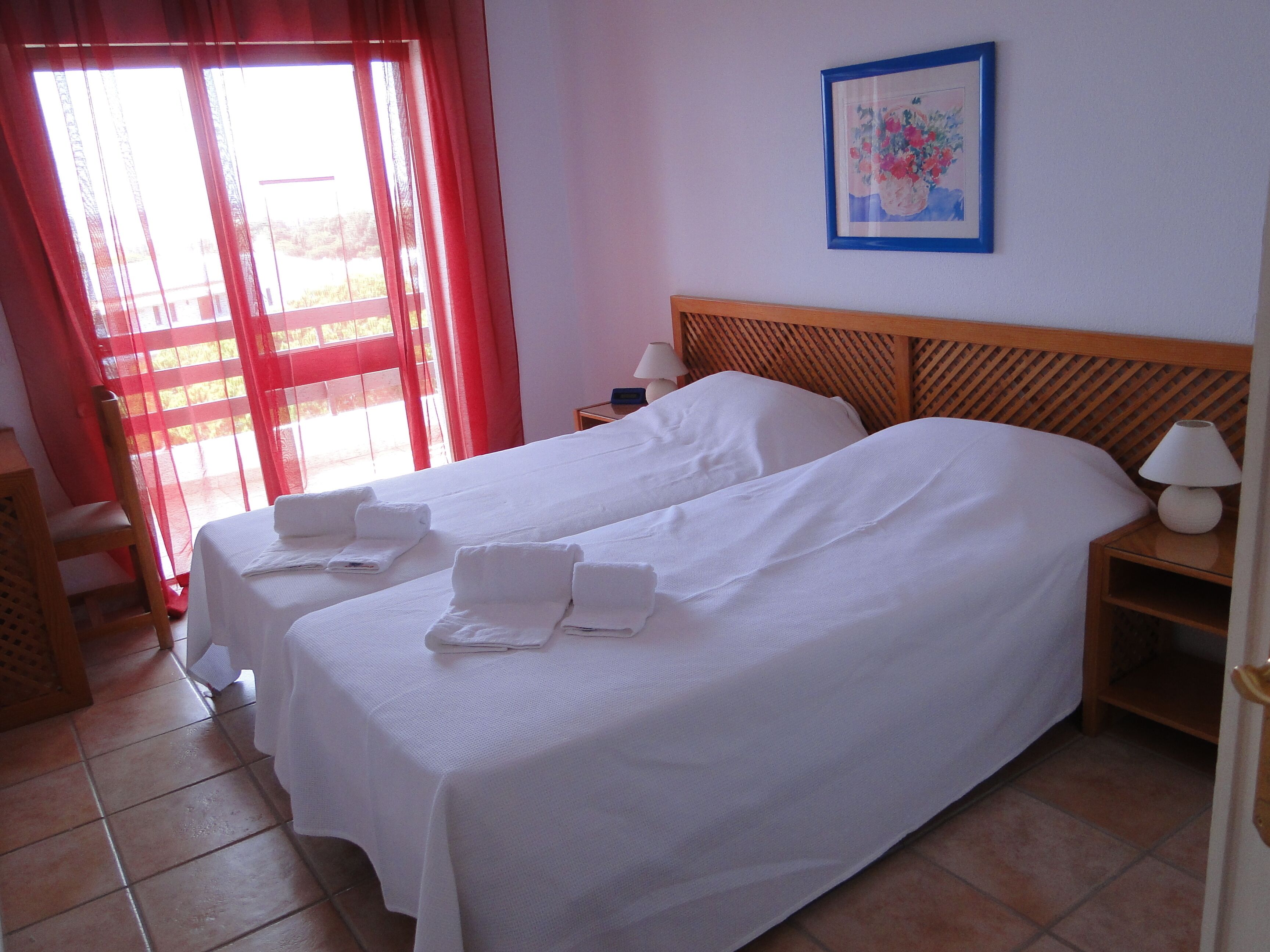 A hotel room with two beds, a wooden headboard, a painting on the wall, and a window with red curtains.
