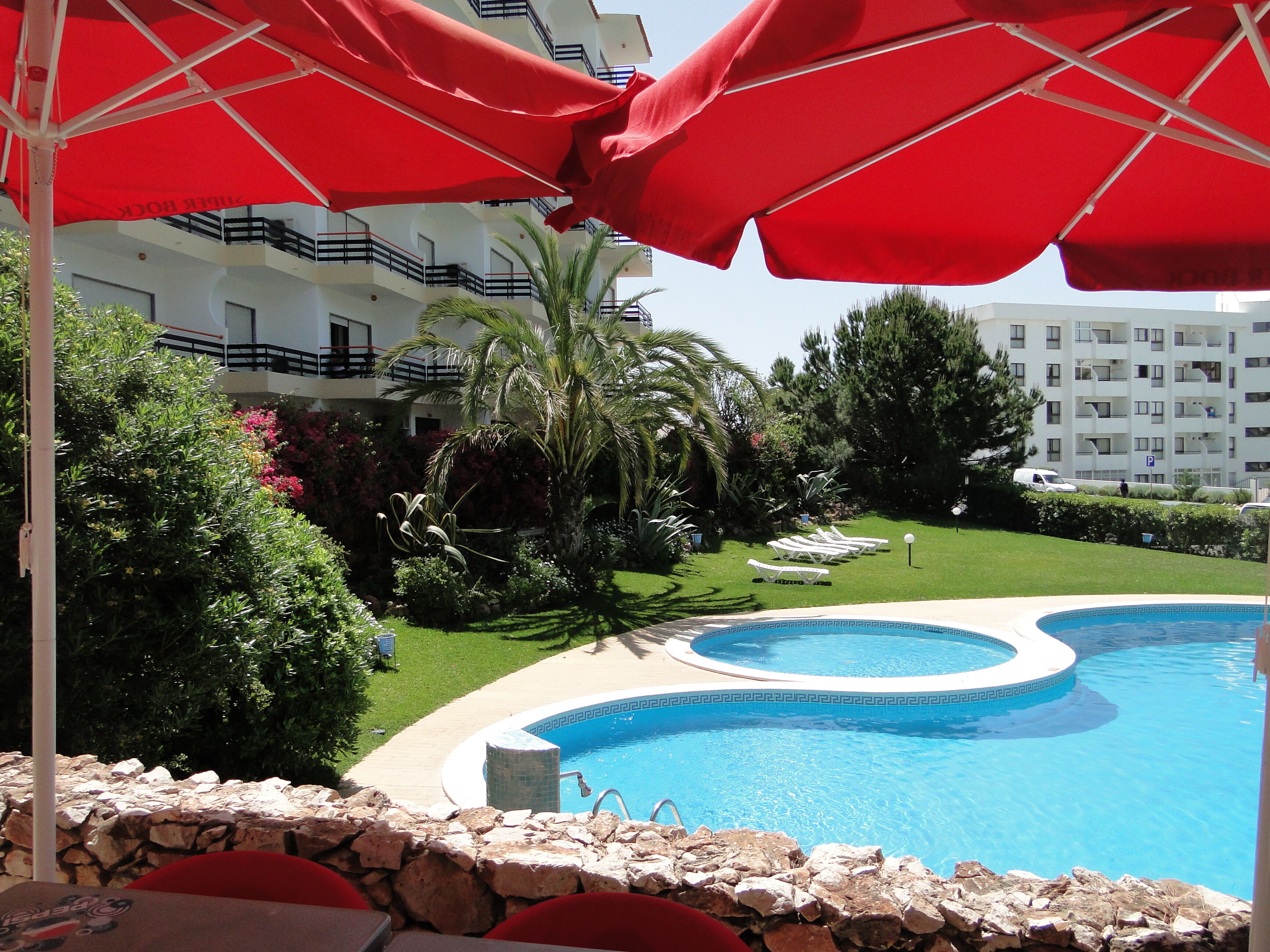 A pool area with a stone wall, red umbrellas, and a building in the background.