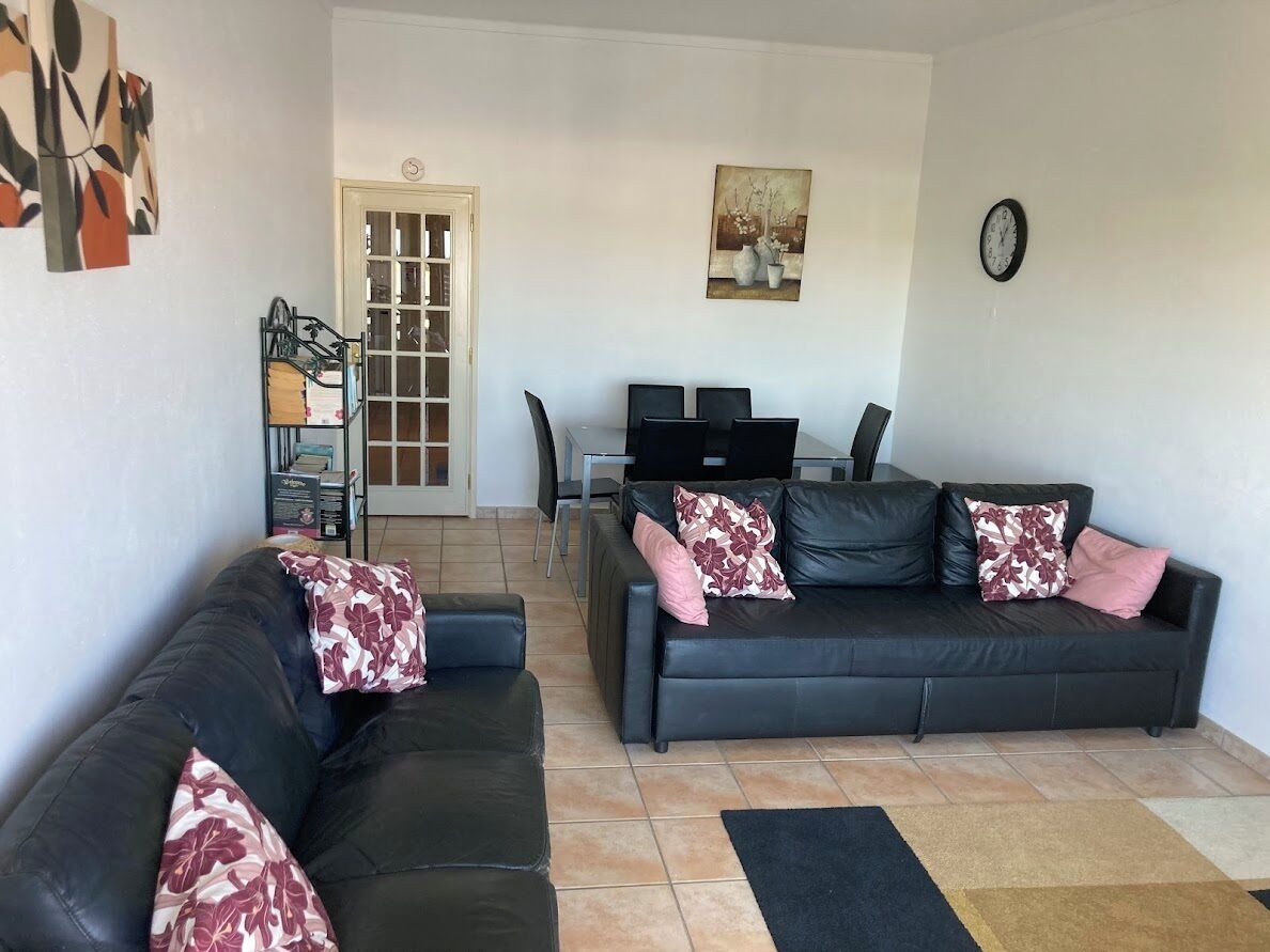 A living room with a black leather sofa, a dining table with chairs, and a clock on the wall.