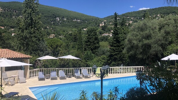 Un espace piscine doté de chaises longues et de parasols, entouré d’une végétation luxuriante et offrant une vue sur une colline parsemée de bâtiments.