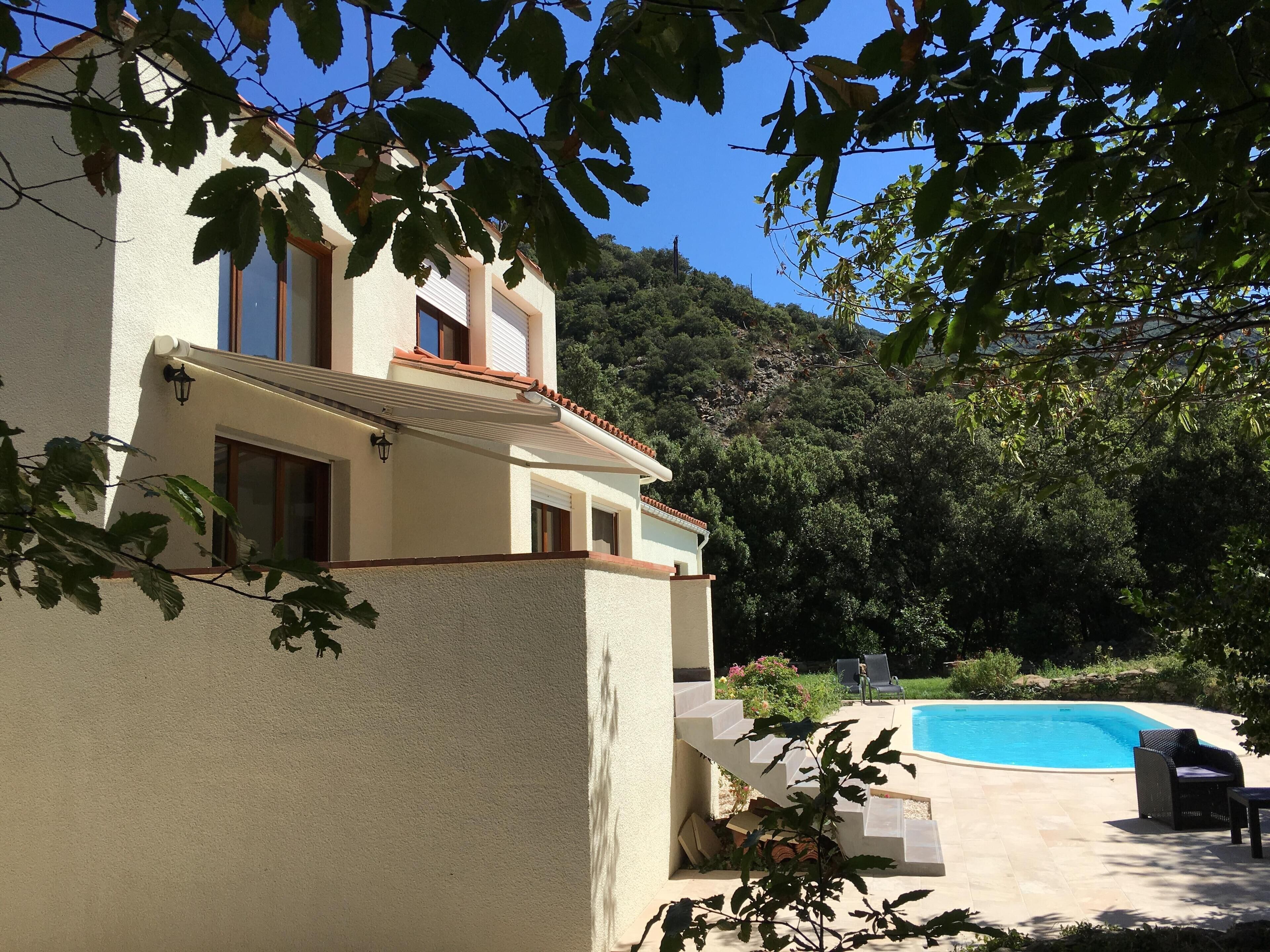 A two-story house with a swimming pool, surrounded by greenery and hills.