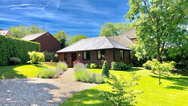 A two-story wooden house with a gravel driveway leading to a front door, surrounded by a well-maintained lawn and various shrubs.