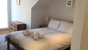 A bedroom with a large bed, a wooden dresser, a nightstand, a floor lamp, and a picture on the wall. - Harbour View Apartment has stunning views of St Ives harbour and beaches. (St Ives)