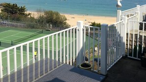 A tennis court with a white fence, a beach with people, and the ocean in the background. - Harbour View Apartment has stunning views of St Ives harbour and beaches. (St Ives)