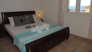 Un lit bien fait, recouvert d’une couvre-lit à rayures, avec une tête de lit en bois et deux oreillers. Une table de chevet avec une lampe et un livre se trouve à côté du lit. On aperçoit une fenêtre avec des rideaux, et un tableau est accroché au mur.