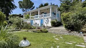 A two-story house with a white exterior and blue shutters, surrounded by a well-maintained garden with a stone pathway leading to the entrance.