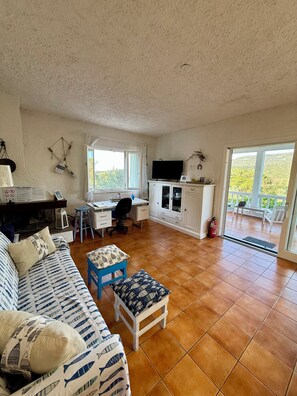 A living room with a sofa, a small table, a desk, and a television. There is a window with curtains, a balcony with a view of greenery, and a fire extinguisher.