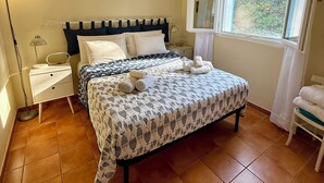 A bedroom with a bed, a nightstand, a lamp, an air conditioning unit, a window with a view, and a chair with towels.