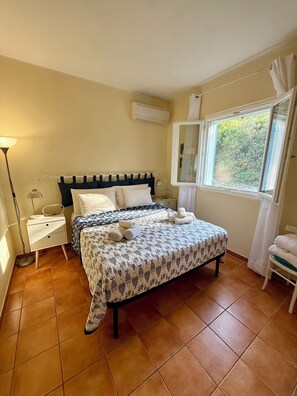 A bedroom with a bed, a nightstand, a lamp, an air conditioner, a window with a view, and a chair.