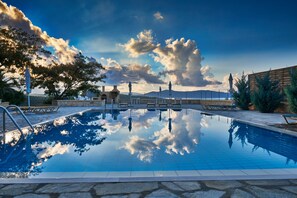 Une piscine aux eaux cristallines, bordée de pierres, avec des bancs et une vue sur les montagnes sous un ciel partiellement nuageux.