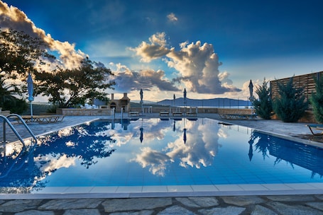 A clear swimming pool with a stone border, benches, and a view of mountains under a partly cloudy sky.