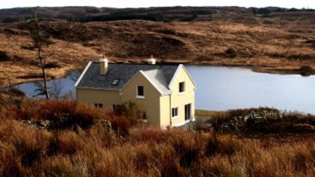 A single-story house with a gabled roof, situated next to a body of water, surrounded by dry, grassy terrain.