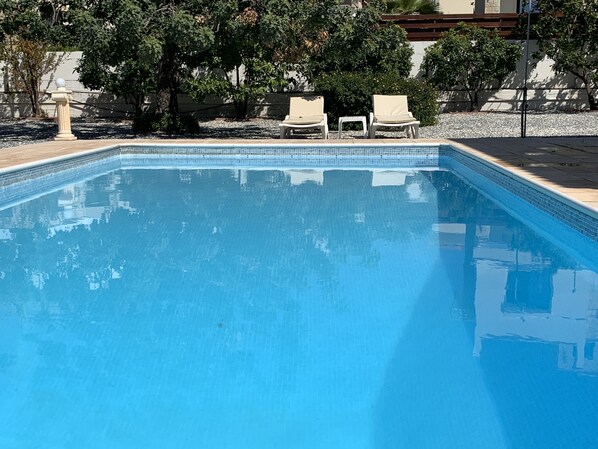A rectangular swimming pool with clear blue water, surrounded by a tiled edge. There are two white lounge chairs and a small table near the pool.