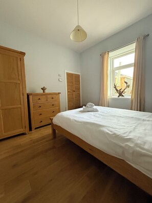 Ein Schlafzimmer mit einem hölzernen Bett, einer Kommode, einem Kleiderschrank, einem Fenster mit Vorhängen und einer Pendelleuchte.